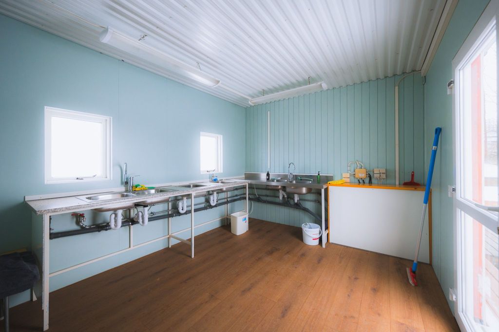 A spacious, minimalist kitchen with light blue walls, countertops, dual sinks, cleaning supplies, and a wooden floor.