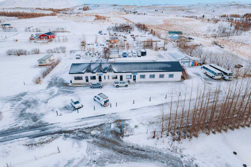 Aerial view of a snowy landscape with a long, single-story building surrounded by parked vehicles. Snow-covered fields and bare trees stretch into the distance.