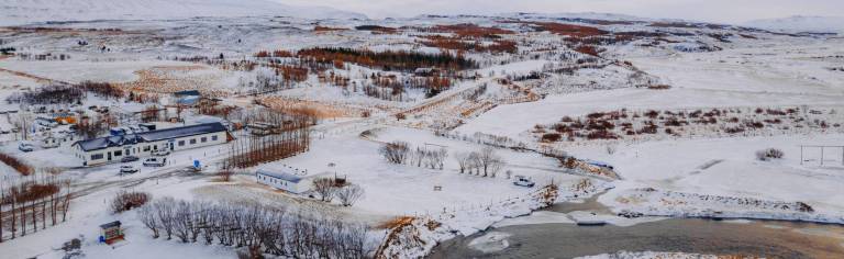 Snowy landscape with scattered buildings near a river, surrounded by barren trees.