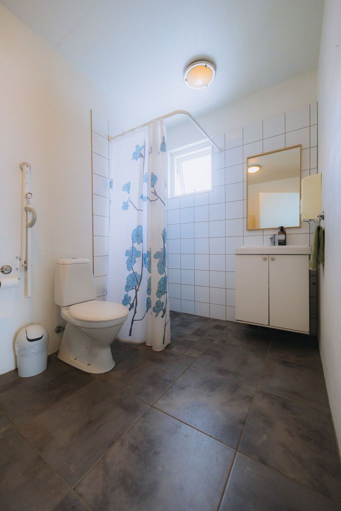 A bright, accessible bathroom with tiled walls and gray flooring. Features a toilet, a blue floral shower curtain, and a sink with a mirror above.