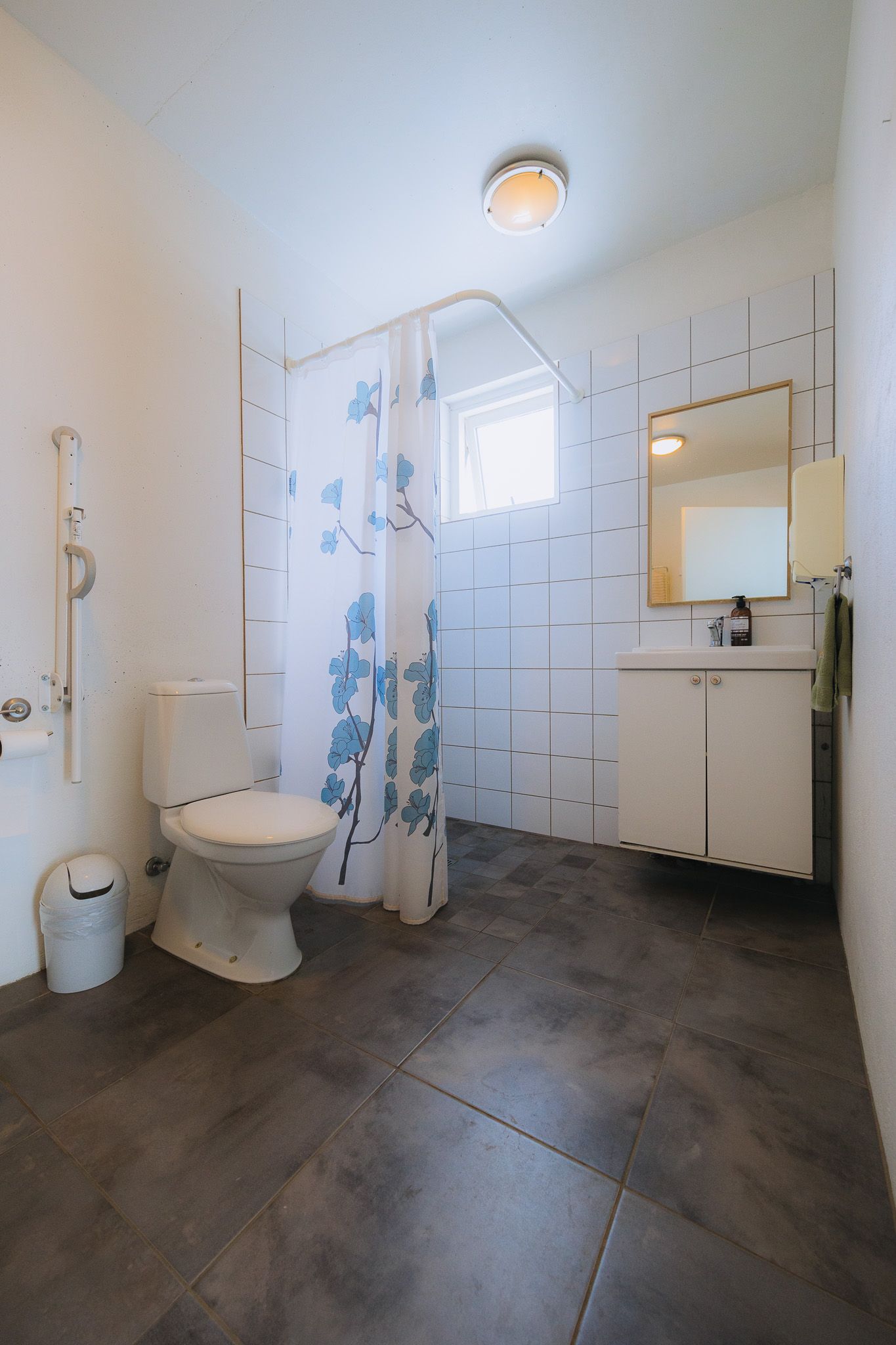 A bright, accessible bathroom with tiled walls and gray flooring. Features a toilet, a blue floral shower curtain, and a sink with a mirror above.