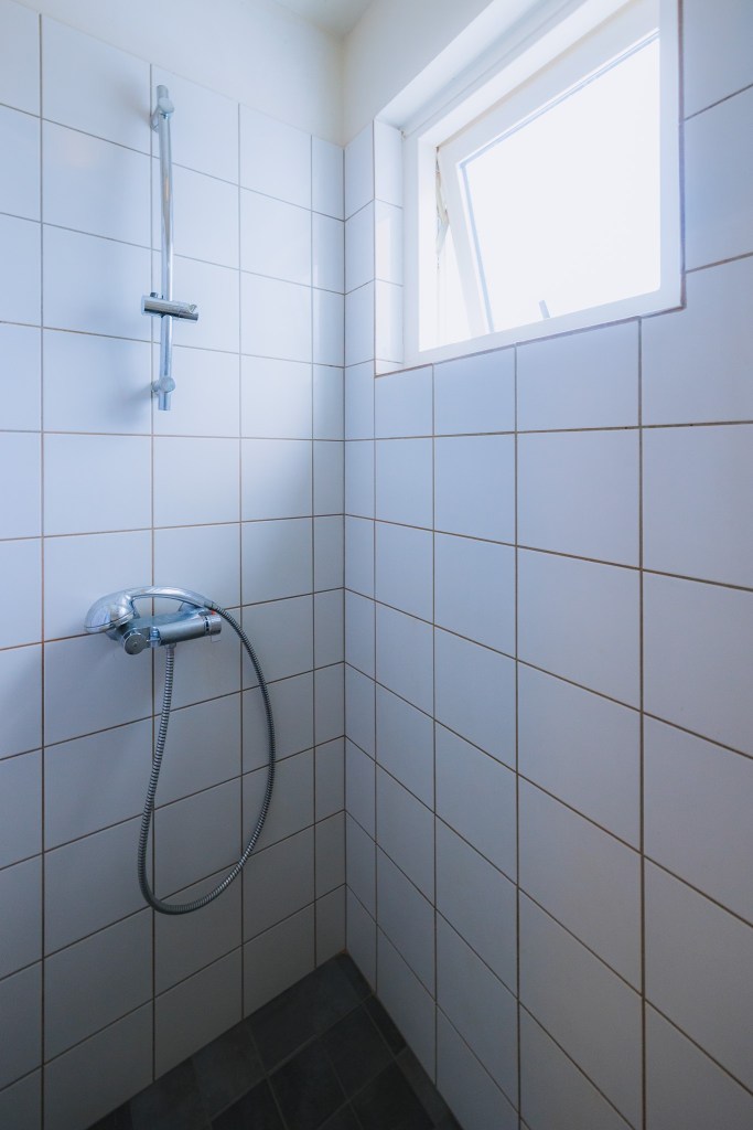 A tiled shower corner with white square tiles, featuring a metal showerhead and hose.