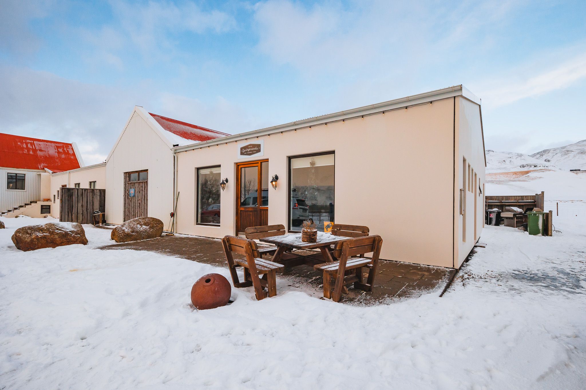 Snow-covered rural scene with a cozy building featuring red roofs and rustic furniture. Bright sky and mountains in the background.