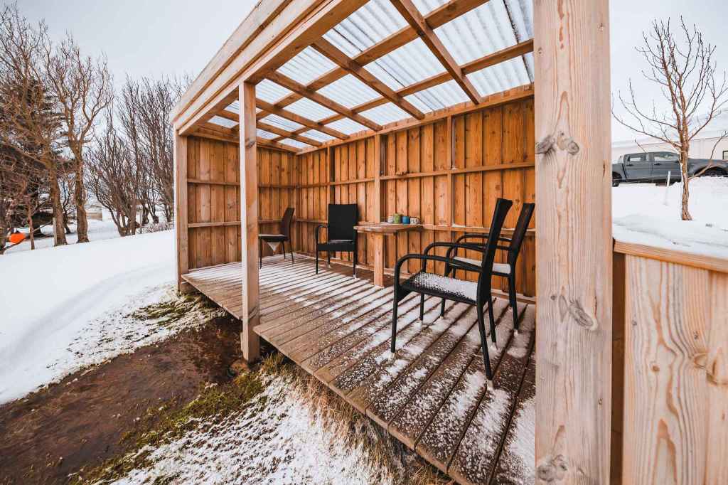 Wooden gazebo with a transparent roof sits in a snowy landscape. Inside, snow-dusted chairs and a small table create a cozy, rustic feel.