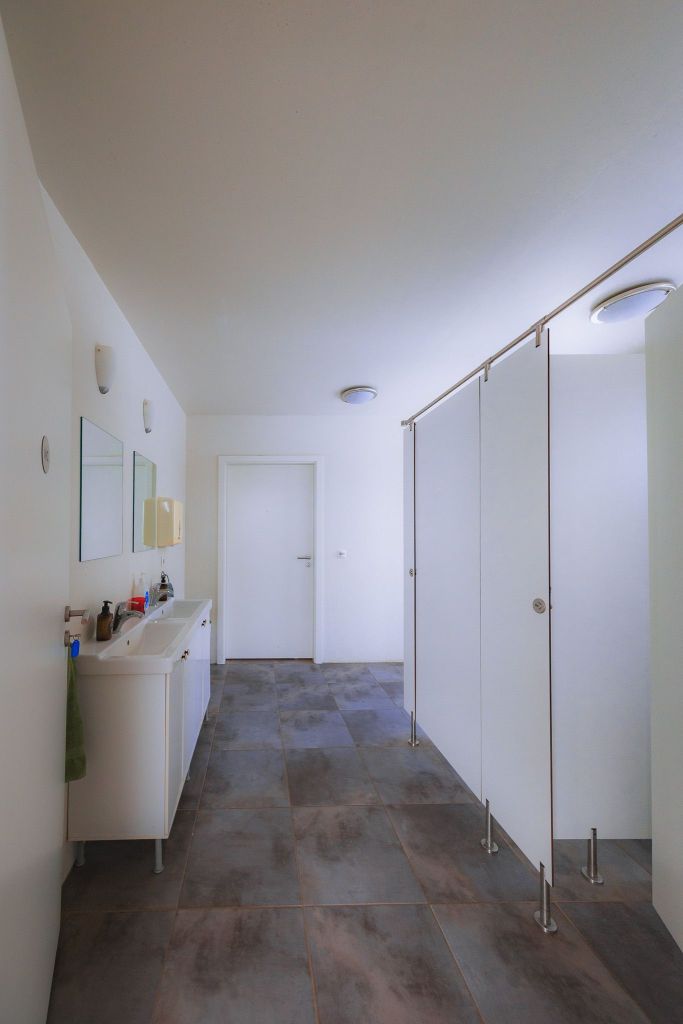 A bright, modern bathroom with large, gray tiles. It features a white double sink on the left and white partitioned stalls on the right.