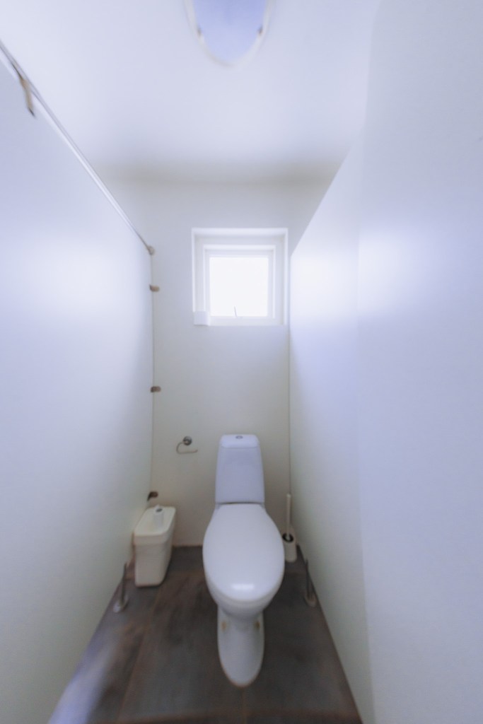 Narrow bathroom with white walls and a single toilet under a small, bright window.