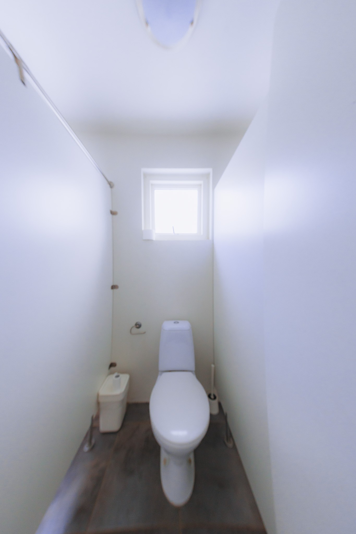 Narrow bathroom with white walls and a single toilet under a small, bright window.