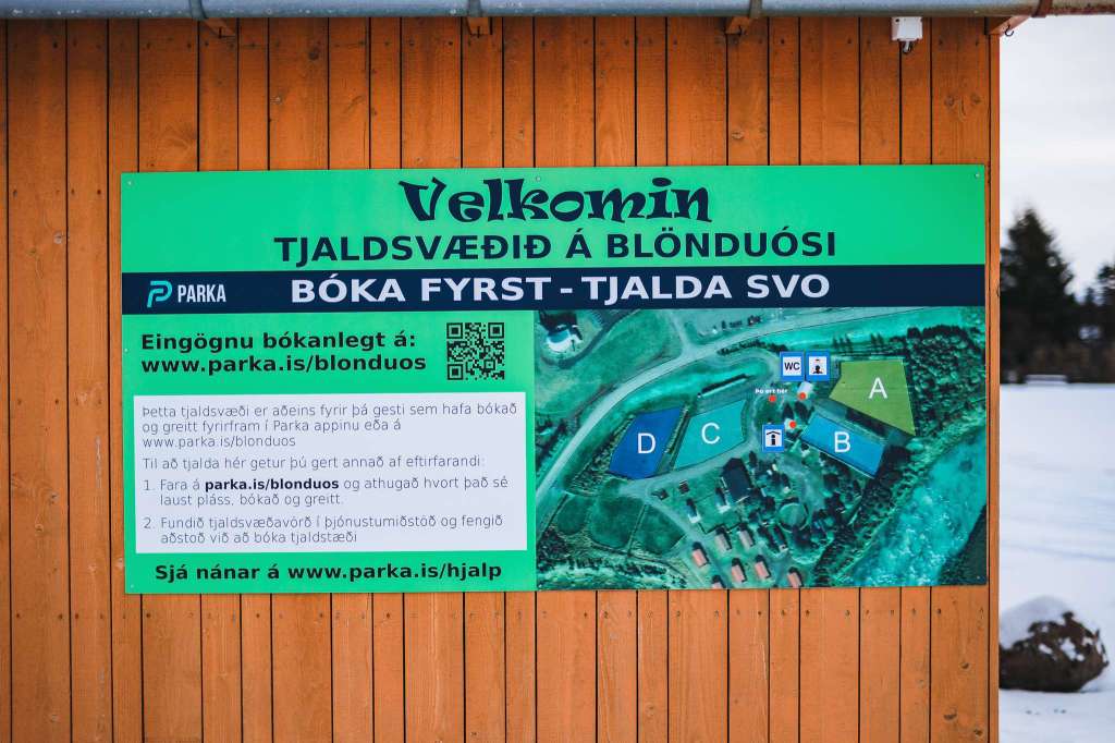 A green and blue sign on a wooden panel welcomes visitors to a campsite in Blönduós, Iceland. It includes a booking website and a campsite map.
