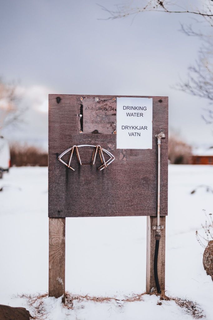 Wooden sign in snowy landscape with a metal water tap and a notice saying "Drinking Water" in English and Icelandic.