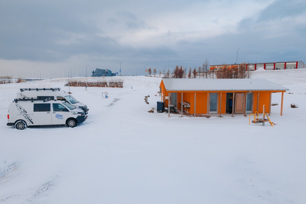 A snow-covered landscape with a bright orange cabin and two white vans parked nearby.
