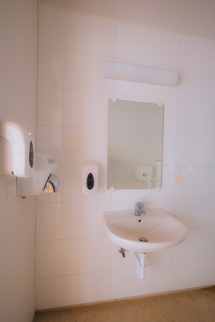 A clean, minimalistic public bathroom with a white sink, mirror, and wall-mounted soap dispenser on tiled walls.