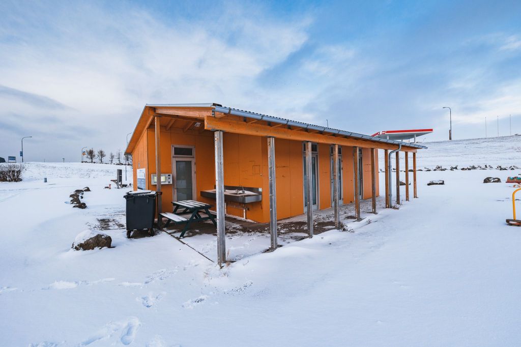 Orange building with wooden posts surrounded by snow, under a cloudy sky. Nearby are benches, a trash can, and distant power lines.