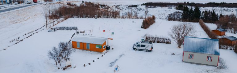 Snowy landscape with two small buildings, one orange and one white, near a parked vehicle. Bare trees and snow-covered fields.