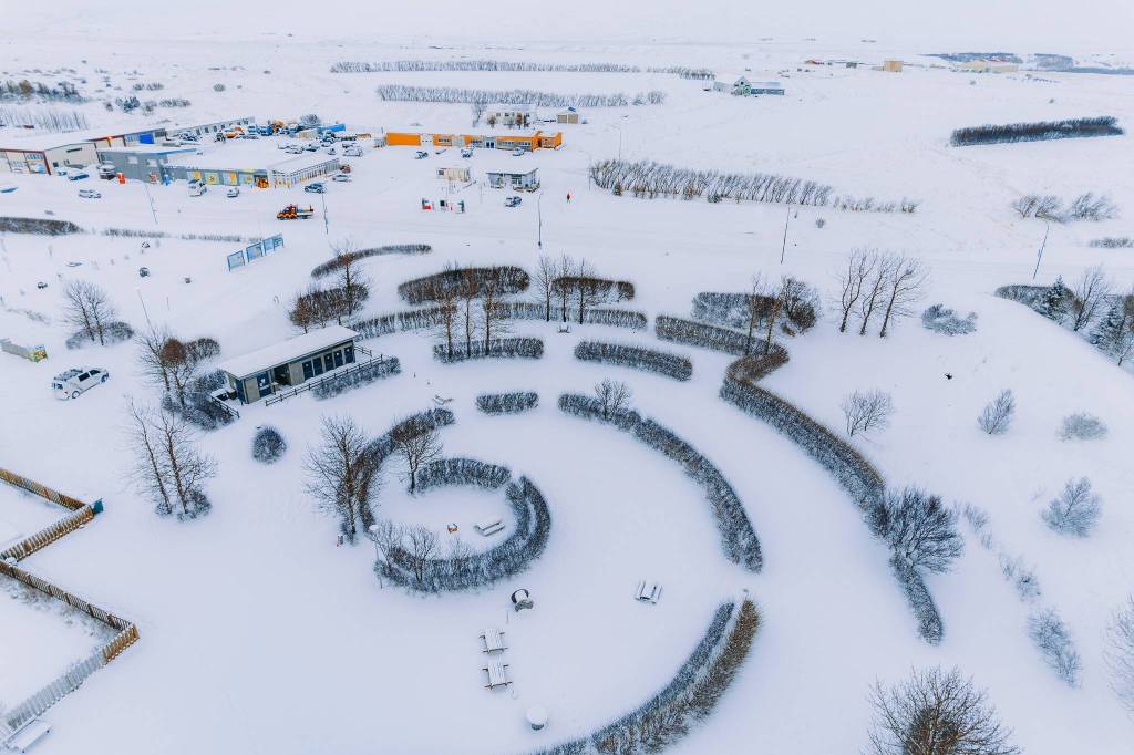 Aerial view of a snow-covered landscape with a spiral-shaped garden in the foreground. Buildings and several snow-dusted trees are visible in the background.