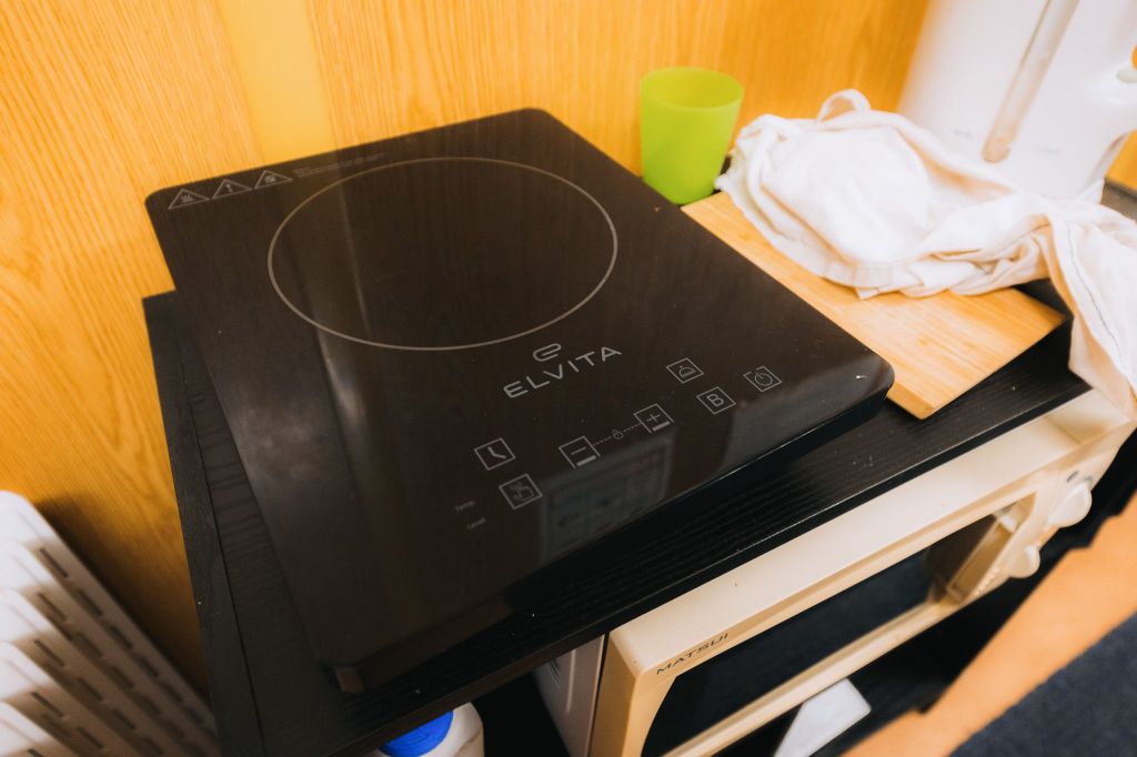 A black Elvita induction cooktop on a counter. Nearby are a folded white cloth, a cutting board, and a green cup, set against a wood-paneled wall.