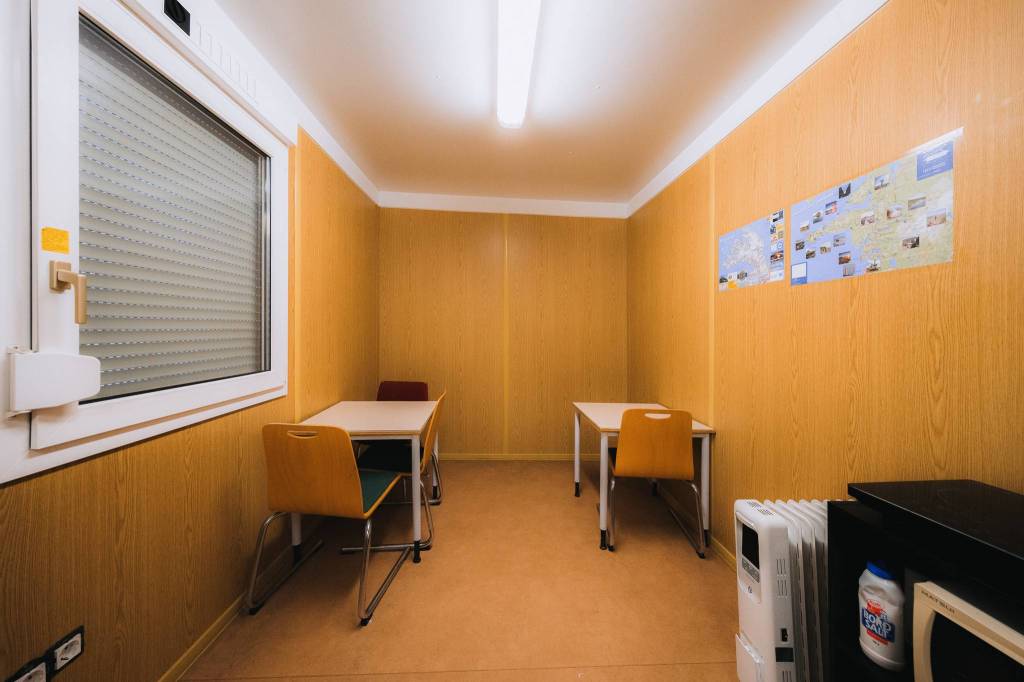 Small, sparsely furnished room with wooden walls, two tables, and mismatched chairs under bright fluorescent lighting. Maps are posted on the wall.