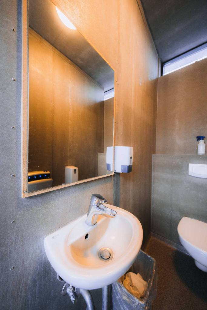 Small bathroom with a wall-mounted sink beneath a mirror. Soap dispenser and trash bin nearby.
