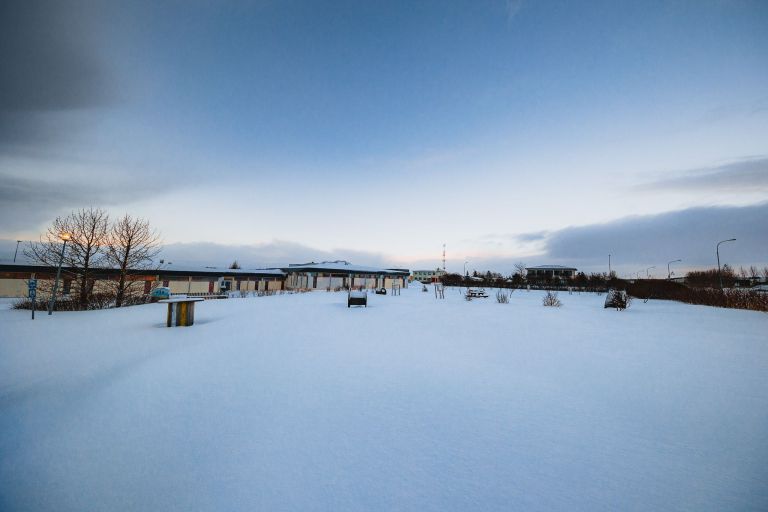 A snow-covered landscape under a clear, blue sky with scattered clouds. In the distance, buildings and leafless trees.