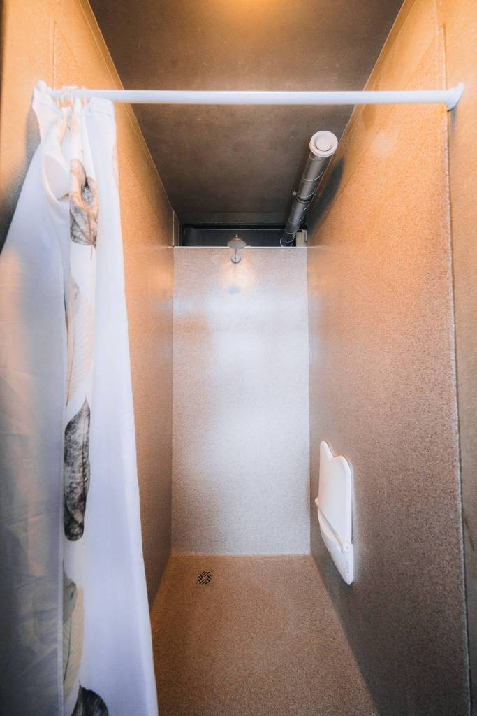 Narrow shower stall with beige walls, a white curtain on the left, and a metal showerhead. A light pipe runs along the ceiling.