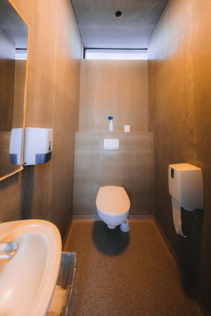 Compact bathroom with a wall-mounted toilet, beige walls, a narrow window, and a small sink on the left. A paper towel dispenser is above the sink.