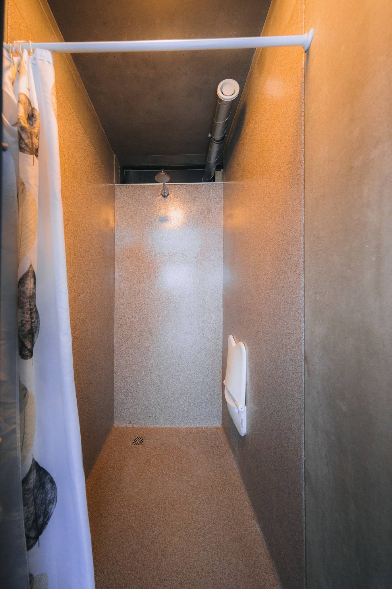 Narrow shower space with beige textured walls, a white plastic foldable bench on the right, ceiling pipe overhead, and a floral curtain on the left.