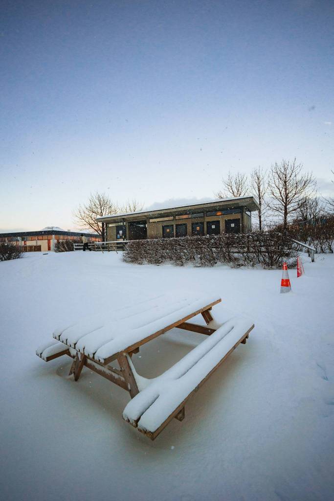 A snow-covered picnic table in a white, wintry landscape, with a small building in the background. The sky is clear, and orange traffic cones are visible nearby.