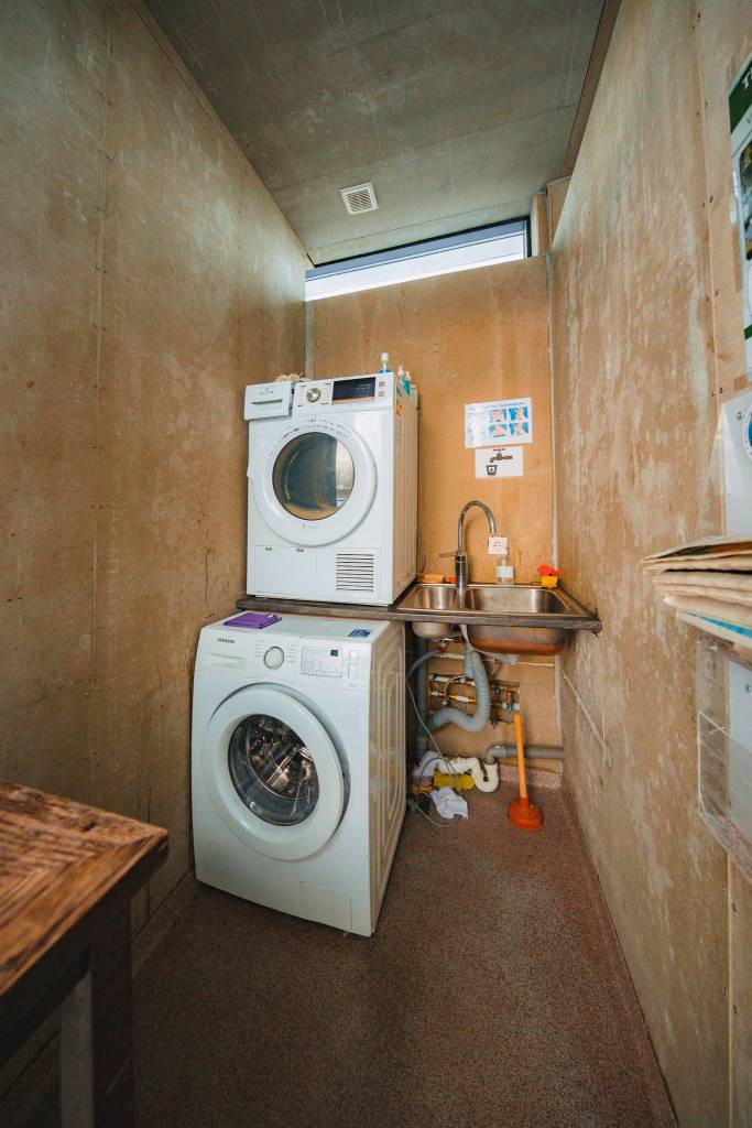Narrow laundry room with stacked washer and dryer on the left, a small stainless steel sink to the right, dim lighting, and basic, unfinished walls.
