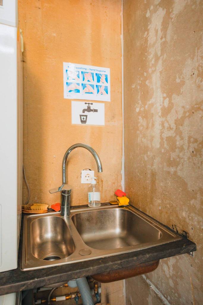 A small, utilitarian sink in a worn, plastered corner. Soap dispenser, sponges, and a brush are on the counter. Handwashing instructions are taped above.