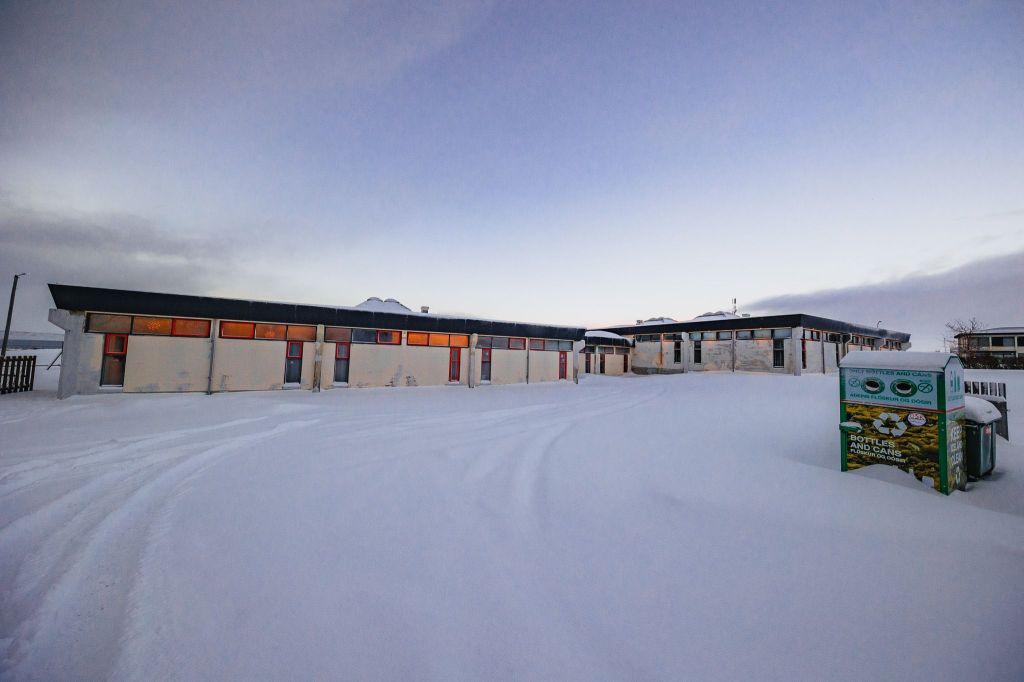 Snow-covered landscape with a long building at dawn. Windows glow with warm light. A green recycling bin is in the foreground.