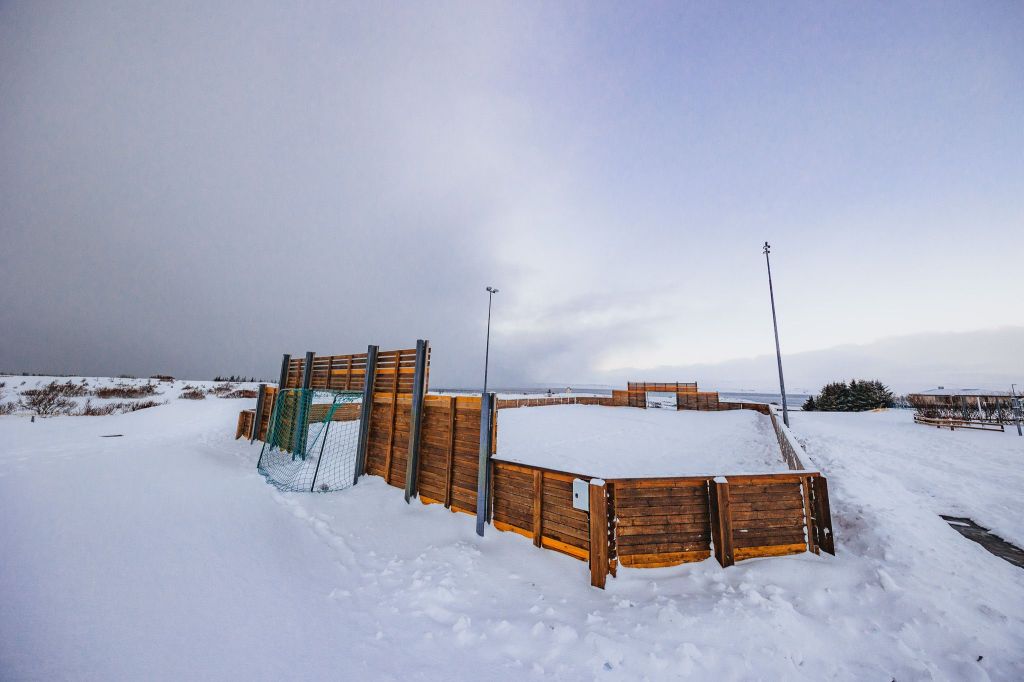 A snowy landscape under a clear sky features a fenced wooden sports field, surrounded by snow. Two tall light poles stand nearby.