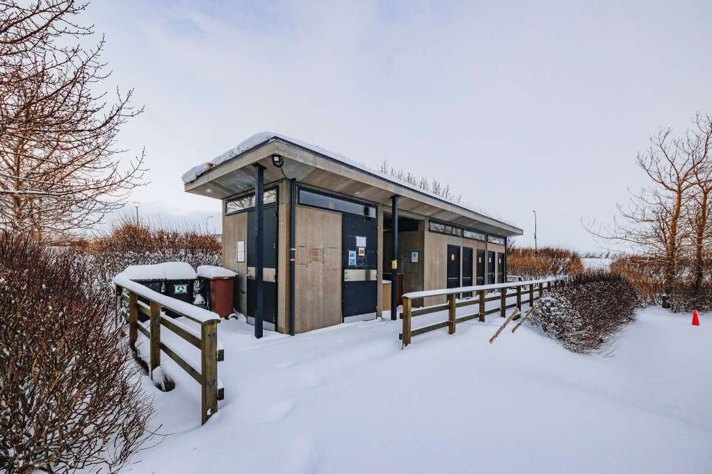 A public restroom facility is surrounded by deep snow, with bare trees and shrubs.