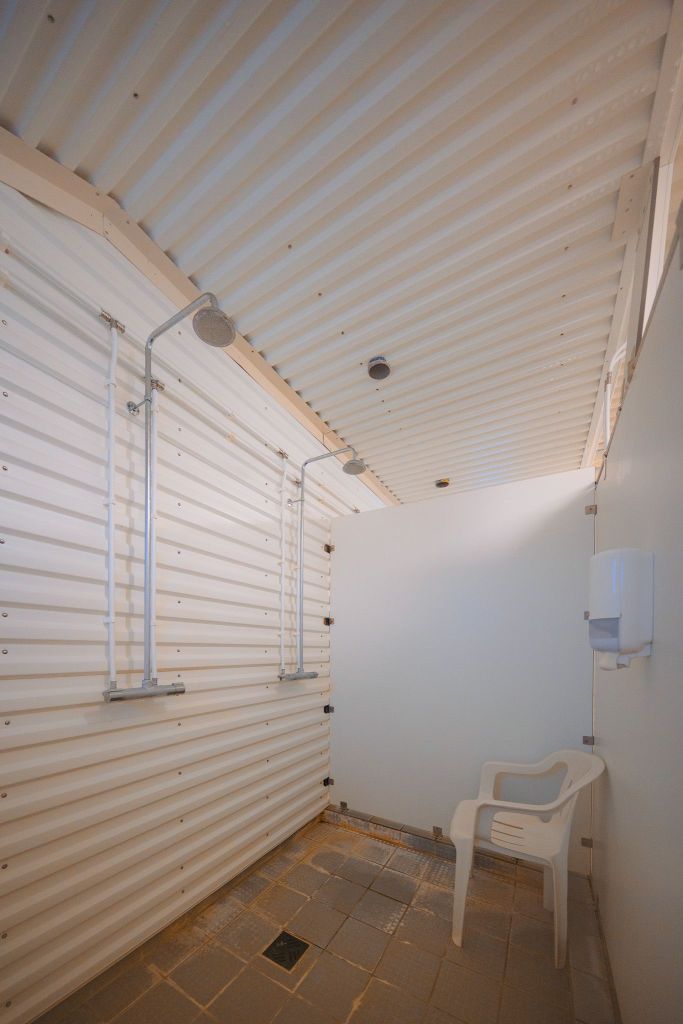 Industrial-style shower room with corrugated metal walls and ceiling. Two showerheads and a white plastic chair are present.