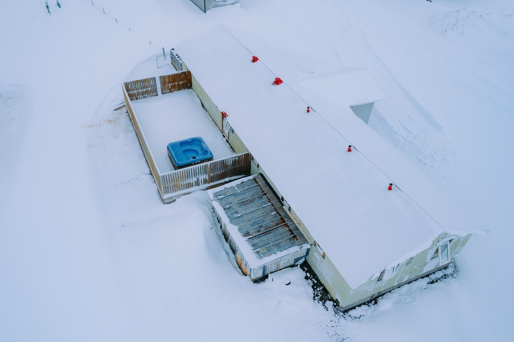 Aerial view of a snow-covered building with red vents. Adjacent is a fenced area with a blue hot tub. Surroundings are blanketed in snow.