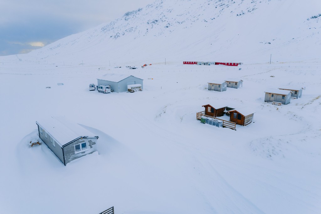 Snow-covered landscape with scattered cabins and a large building. A mountain looms in the background under a cloudy sky.