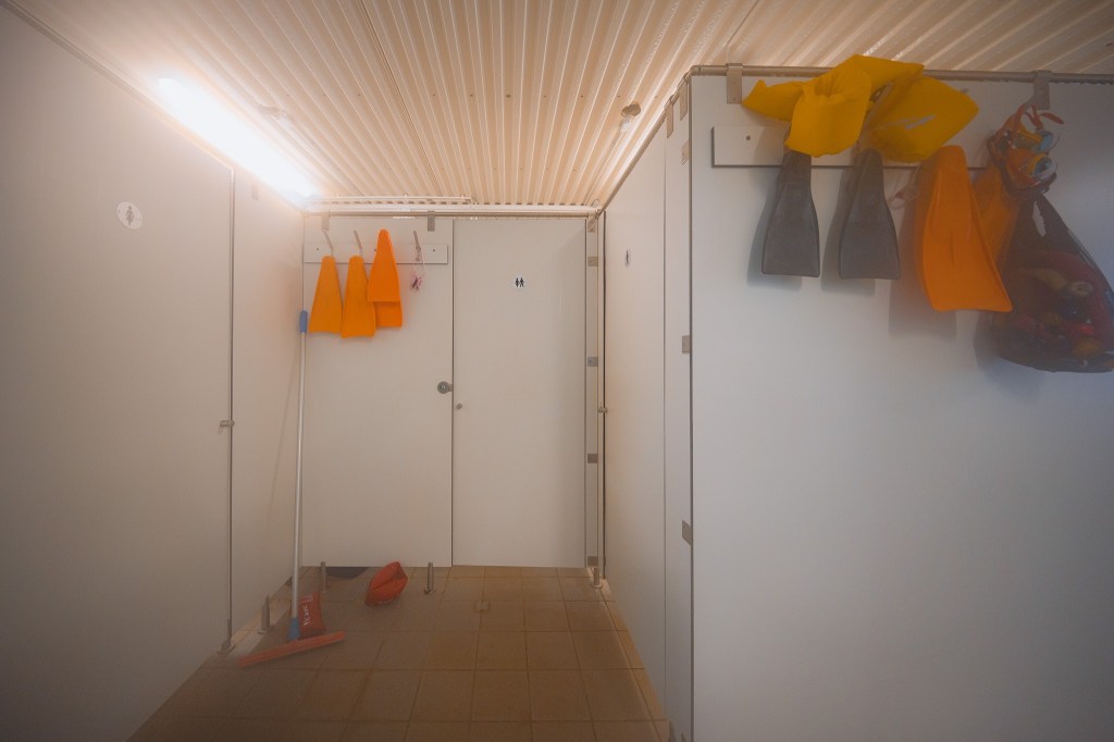 Indoor pool changing area with white walls and ceiling. Bright light overhead. Orange life vests and flippers hang on wall hooks.