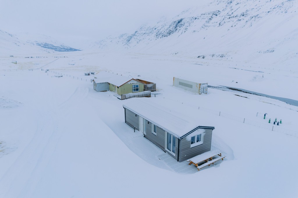 Snow-covered cabins in a vast, tranquil valley surrounded by snowy mountains. The scene is serene and remote, with pale blue skies above.