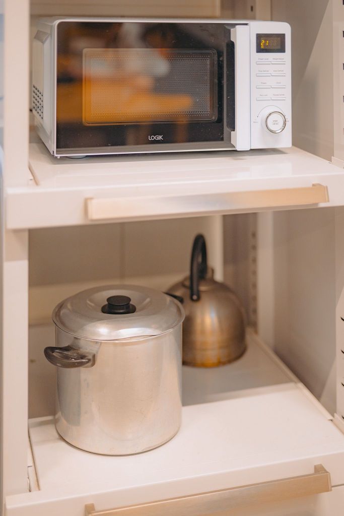 White microwave on a shelf above a metal pot and brass kettle on a lower shelf in a kitchen.