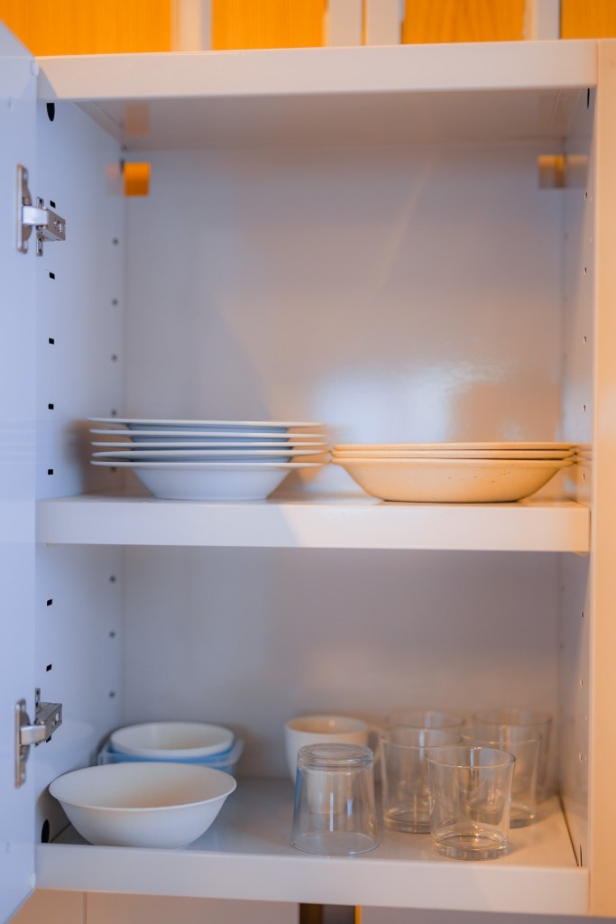 Open kitchen cabinet with neatly stacked plates on the top shelf and bowls with glasses on the bottom.