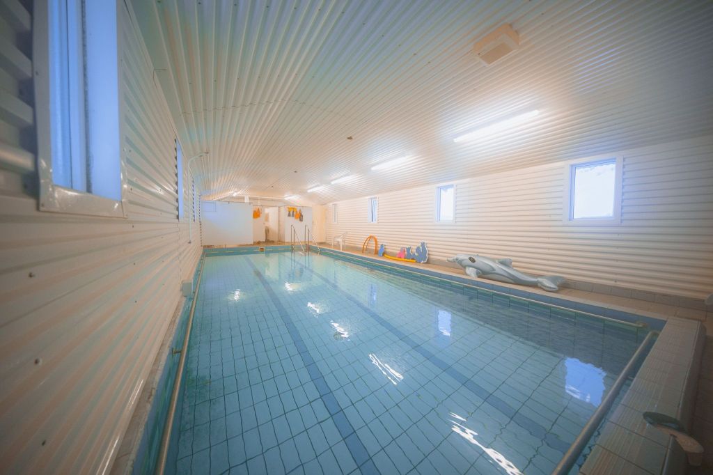 Indoor swimming pool with calm, clear water reflecting ceiling lights. White paneled walls and small windows create a serene, bright atmosphere. Pool toys are visible at the far end.