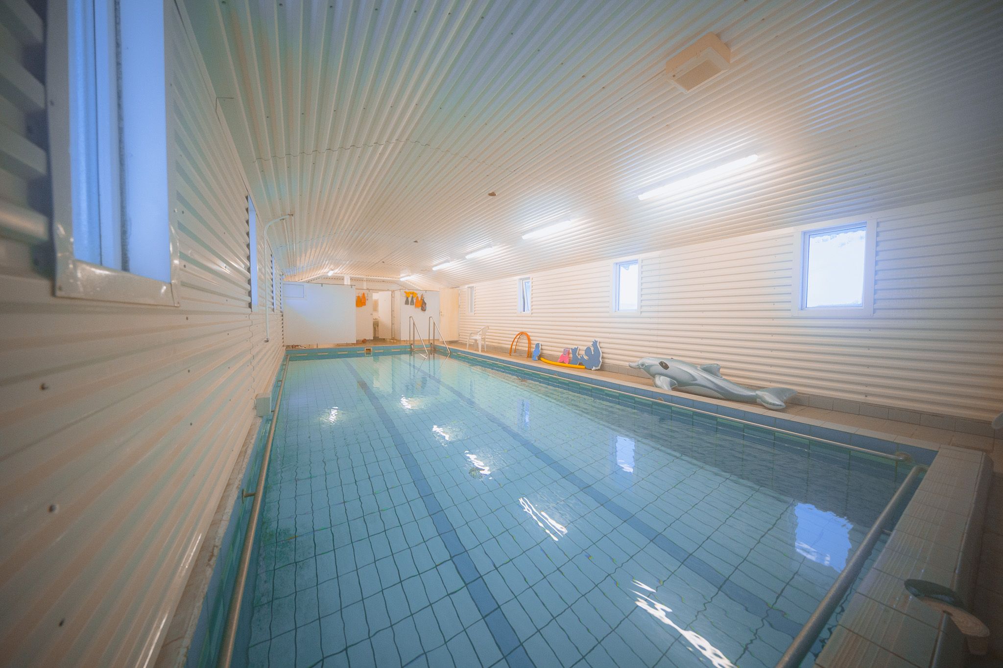 Indoor swimming pool with calm, clear water reflecting ceiling lights. White paneled walls and small windows create a serene, bright atmosphere. Pool toys are visible at the far end.