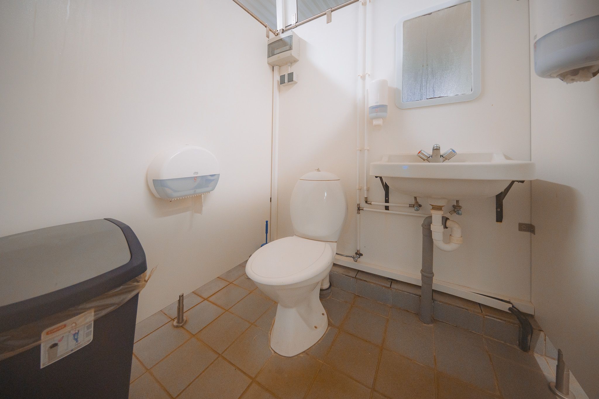 Small, clean bathroom with white walls and gray-tiled floor. Features a toilet, wall-mounted sink, mirror, and a waste bin.