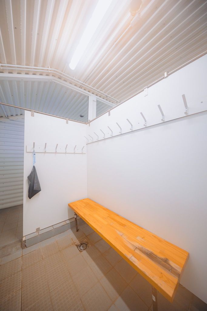 Empty locker room with white walls, wooden bench, and metal hooks. A single plastic bag hangs on a hook.