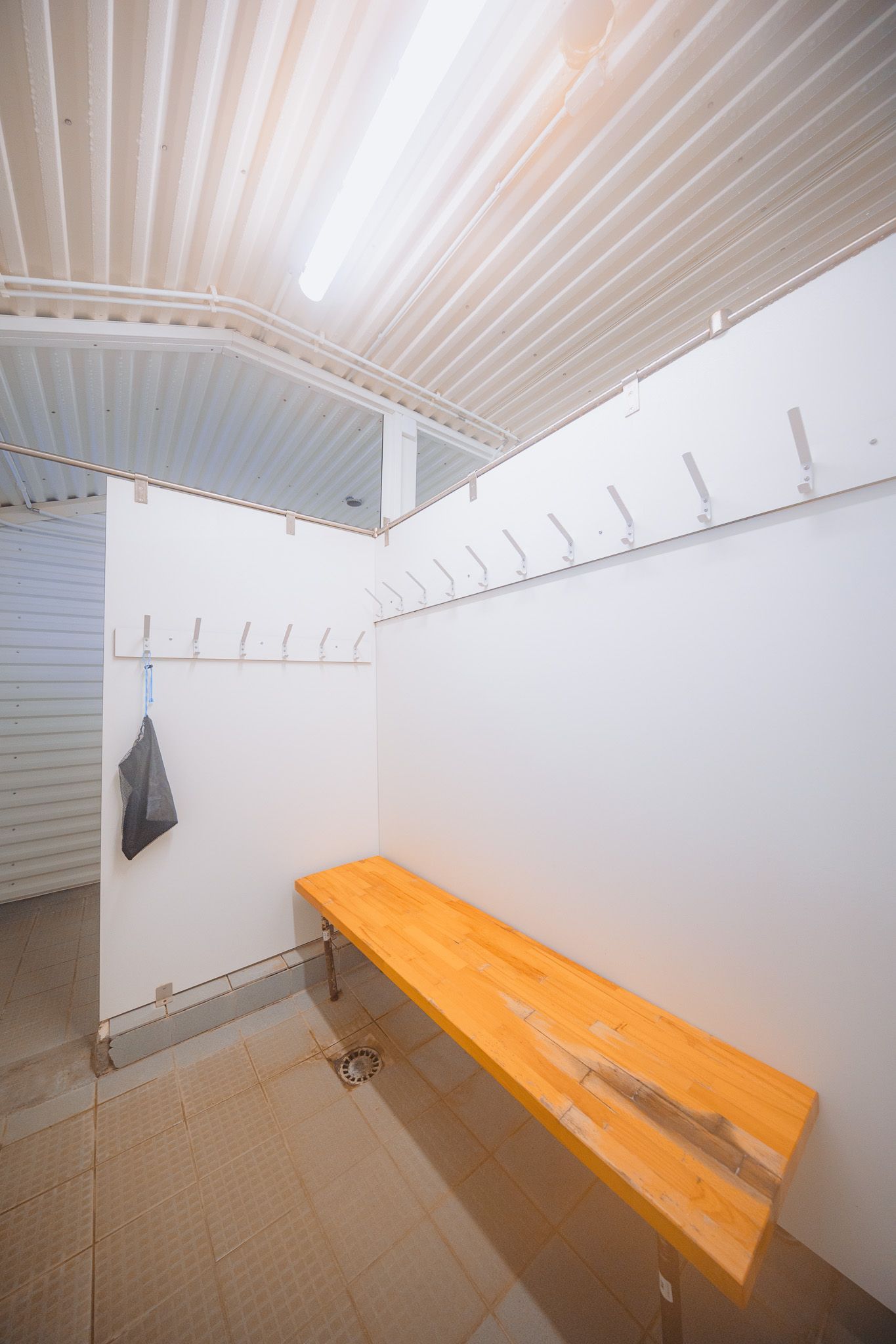 Empty locker room with white walls, wooden bench, and metal hooks. A single plastic bag hangs on a hook.