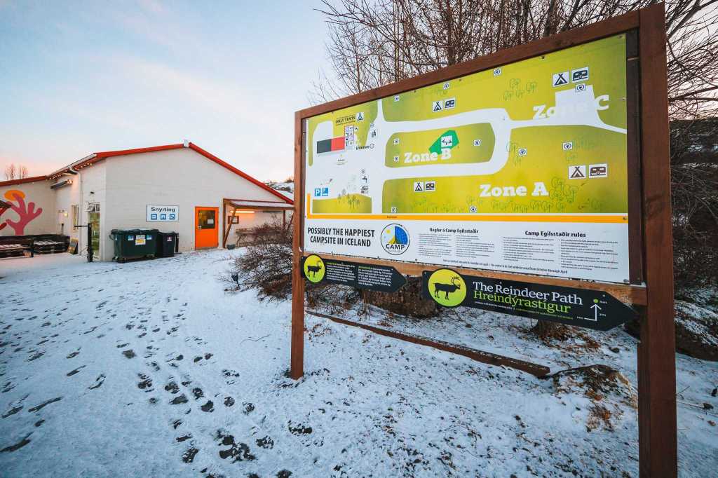 A snowy campsite entrance with a large map sign titled “Possibly the Happiest Campsite in Iceland.” Snowy path, building with red trim, and bins beside it.