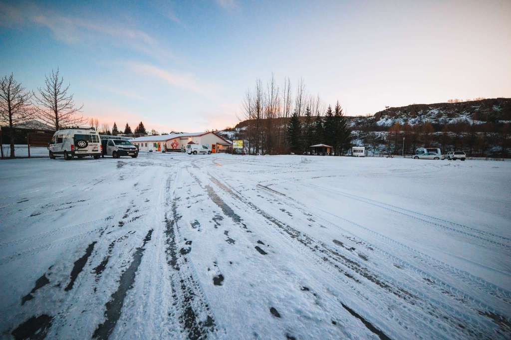 Snow-covered parking lot with car tracks leading to a building surrounded by bare trees and hills at dawn. Campers parked to the left.