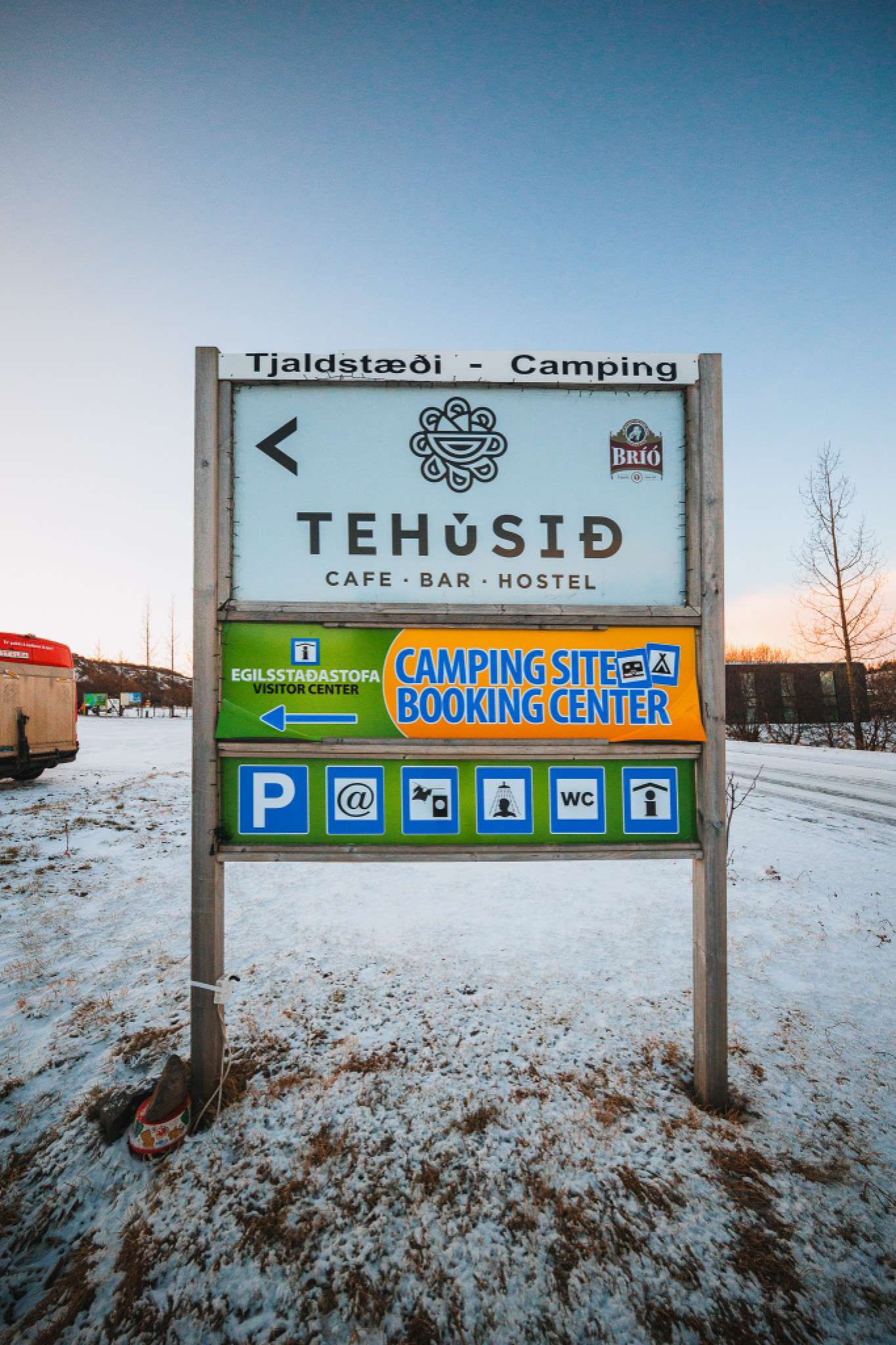 Sign for Tehúsið cafe, bar, hostel, and camping site in snowy landscape. Includes booking center info and icons for parking, restrooms, and amenities.