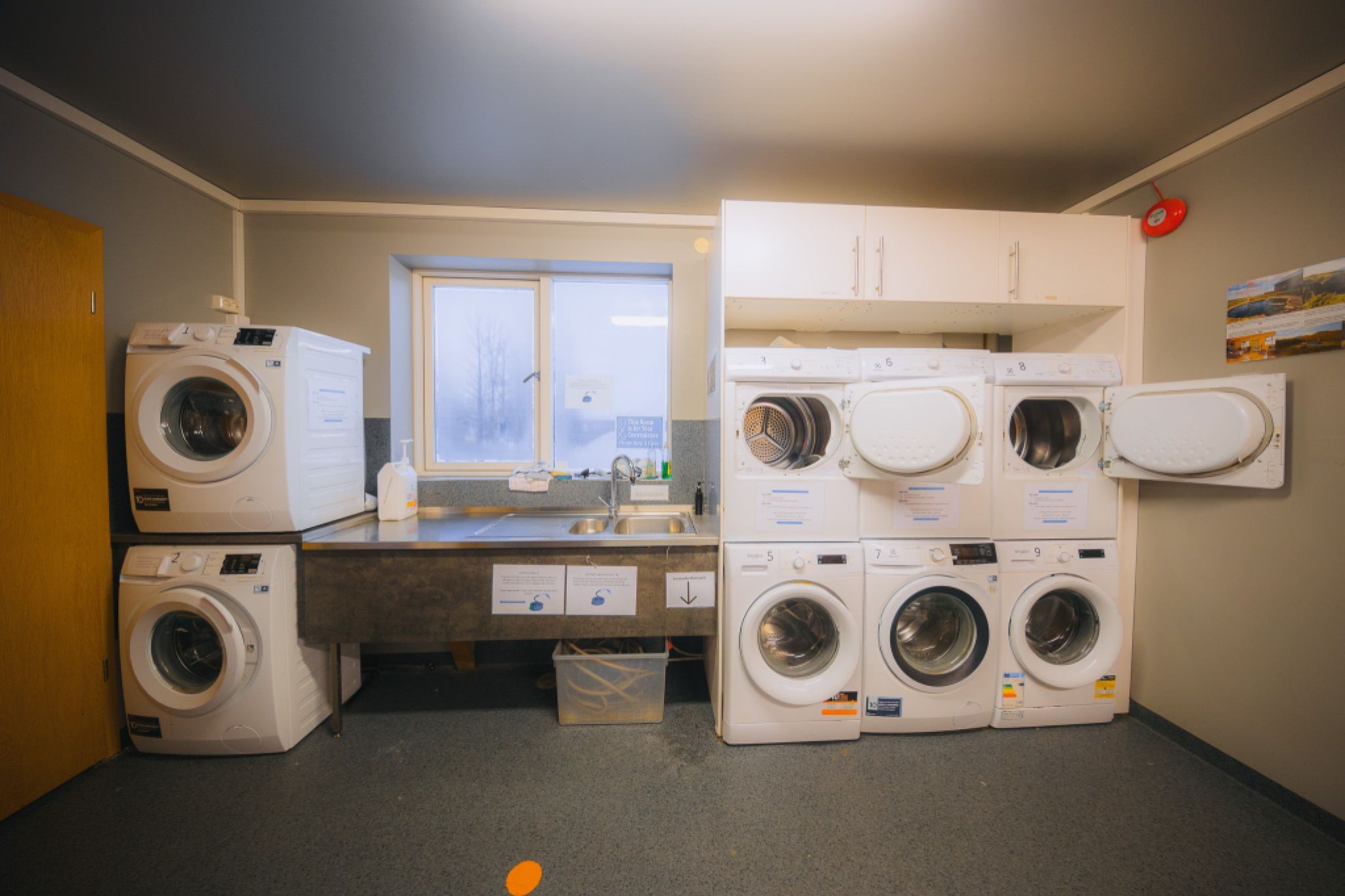 Laundry room with four white washing machines and dryers stacked in two columns. A countertop with a sink, cleaning supplies, and a window above.