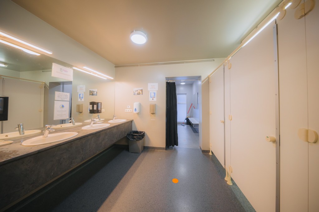 A clean, well-lit bathroom with beige stalls, a long counter with three sinks, wall-mounted soaps, a black trash bin, and an open door leading to a changing area.