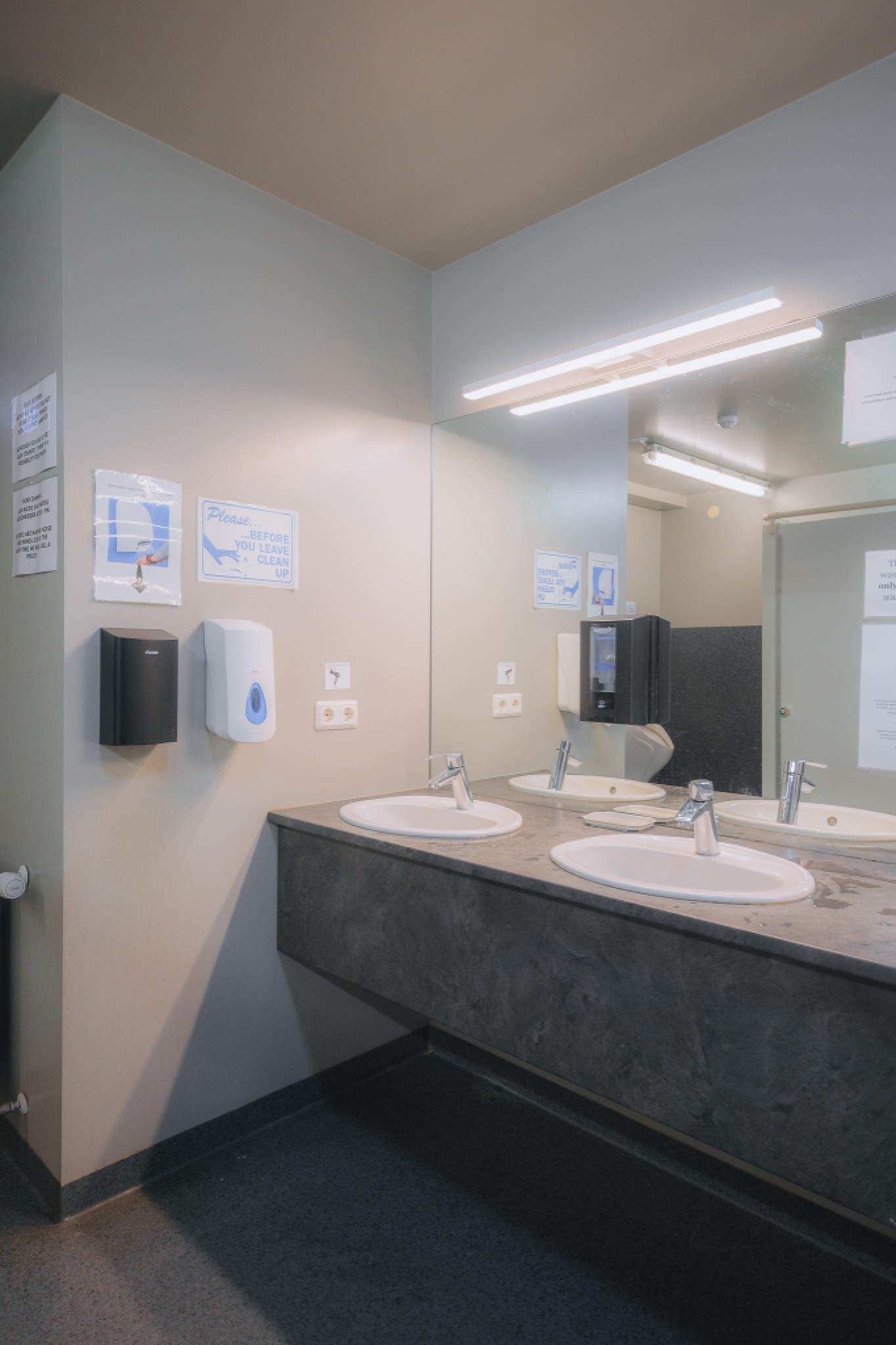 Public restroom with two sinks on a gray countertop, a large mirror above, a dispenser, and signs on the wall.