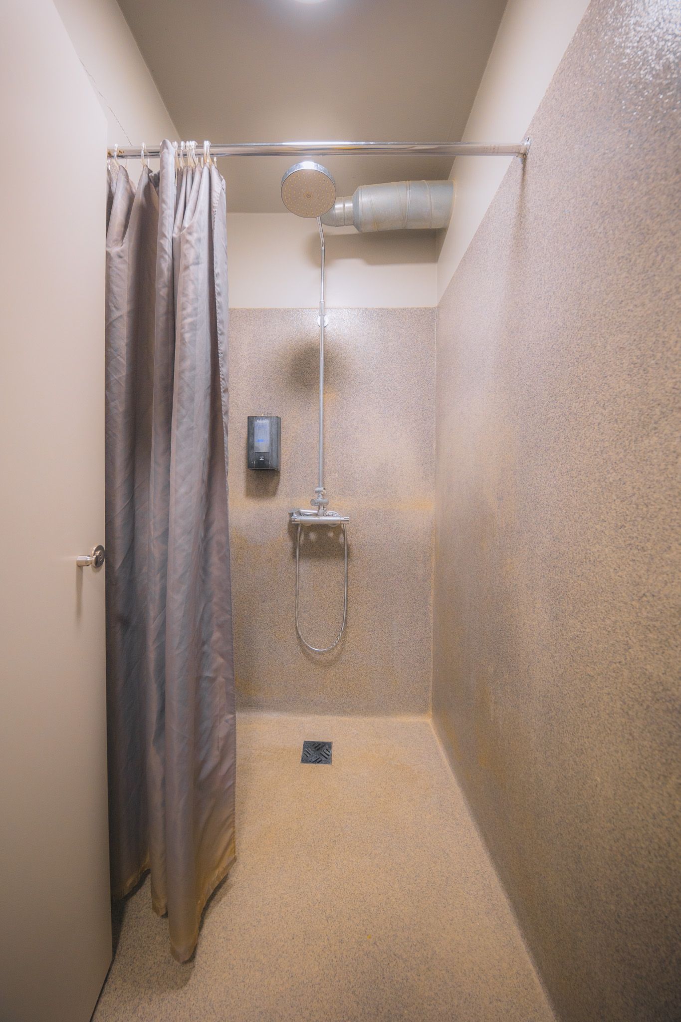 Minimalistic shower room with beige textured walls, ceiling-mounted showerhead, soap dispenser, and a closed gray curtain.